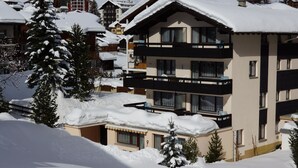 Ein mehrstöckiges Gebäude mit Balkonen, umgeben von schneebedeckten Bäumen und einer Skipiste im Hintergrund.