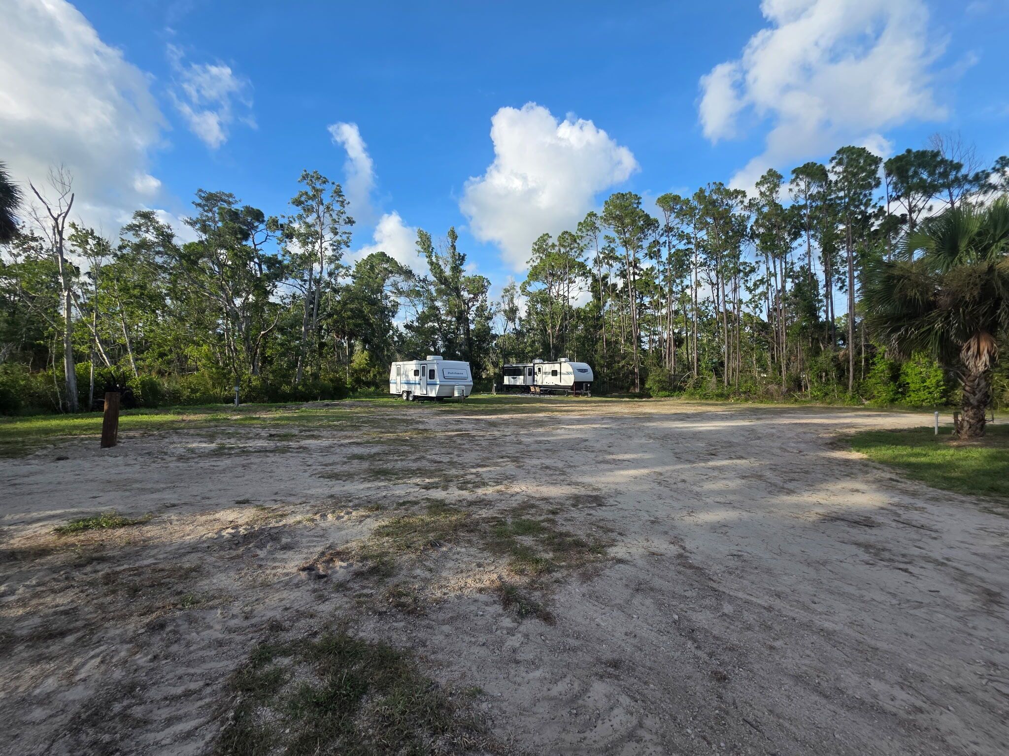 A campsite with two RVs parked on a dirt area, surrounded by trees and greenery.