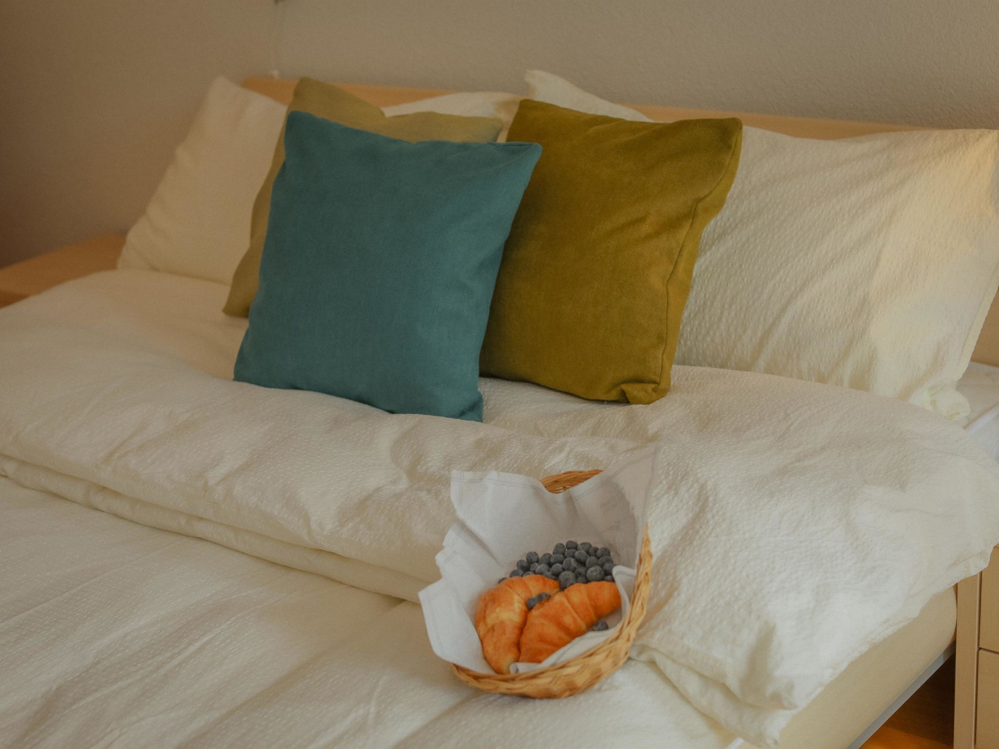 Un lit avec une literie blanche, des oreillers bleu sarcelle et vert olive, et un panier contenant un croissant et des myrtilles.