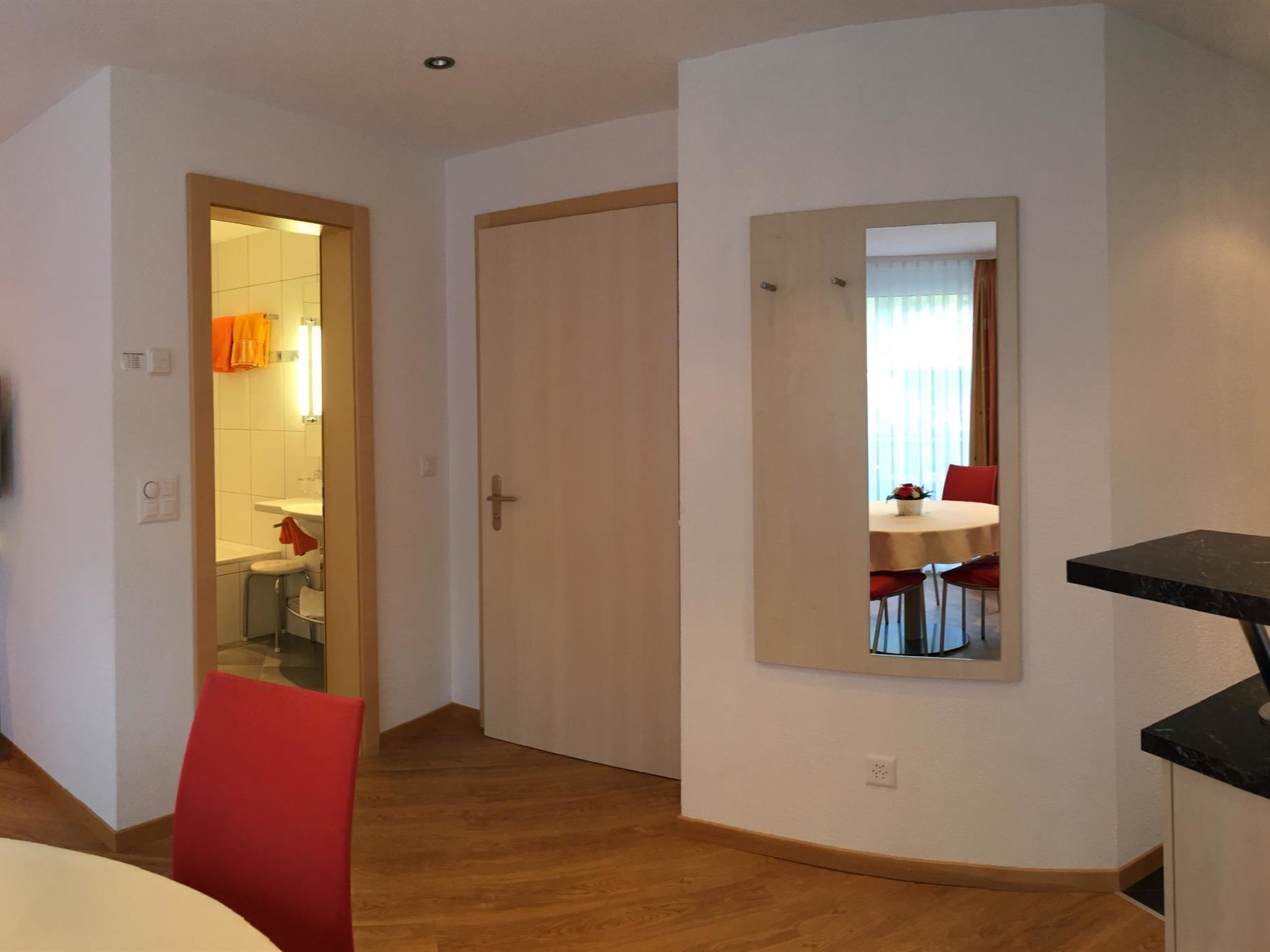 A hotel room with a wooden floor, a red chair, a dining table with red chairs, a mirror, and a door leading to a bathroom.