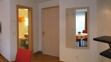 A hotel room with a wooden floor, a red chair, a dining table with red chairs, a mirror, and a door leading to a bathroom.