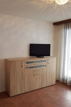 Ein Zimmer mit einem Holzkasten, einem Flachbildfernseher darauf und einem Fenster mit Vorhängen.