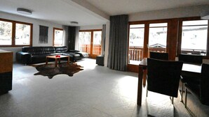 En rummelig opholdsstue med en sort lædersofa, et sofabord og et spisebord med stole. Store vinduer giver naturligt lys og udsigt udad.