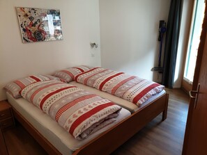 Una cama individual con una colcha estampada y almohadas, una mesita de noche de madera, un espejo montado en la pared y una ventana con cortinas.