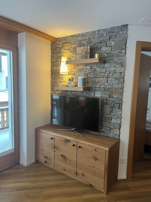 A room with a stone wall, a flat-screen TV on a wooden cabinet, a lamp, and a clock. - Achat Appartement/Fewo 4-Bettwohnung Nr. 3 by Interhome (Saas-Fee)