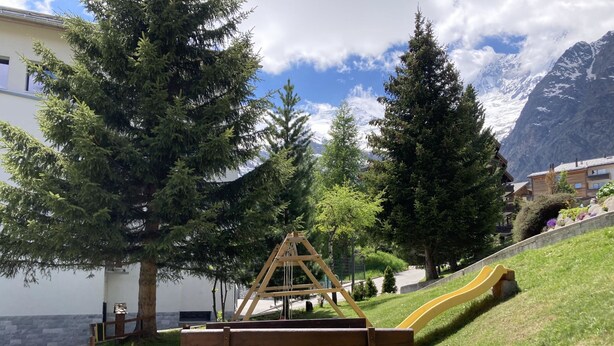 Un parque con un banco de madera, un área de juegos con un tobogán y montañas al fondo.