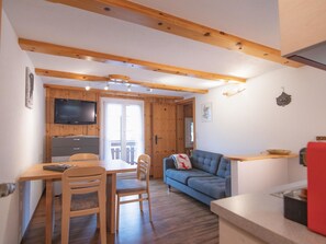 Ein gemütlicher Wohnbereich mit Holzbalkendecke, einem an der Wand montierten Fernseher, einer Couch, einem Esstisch mit Stühlen und einer Küchenzeile.