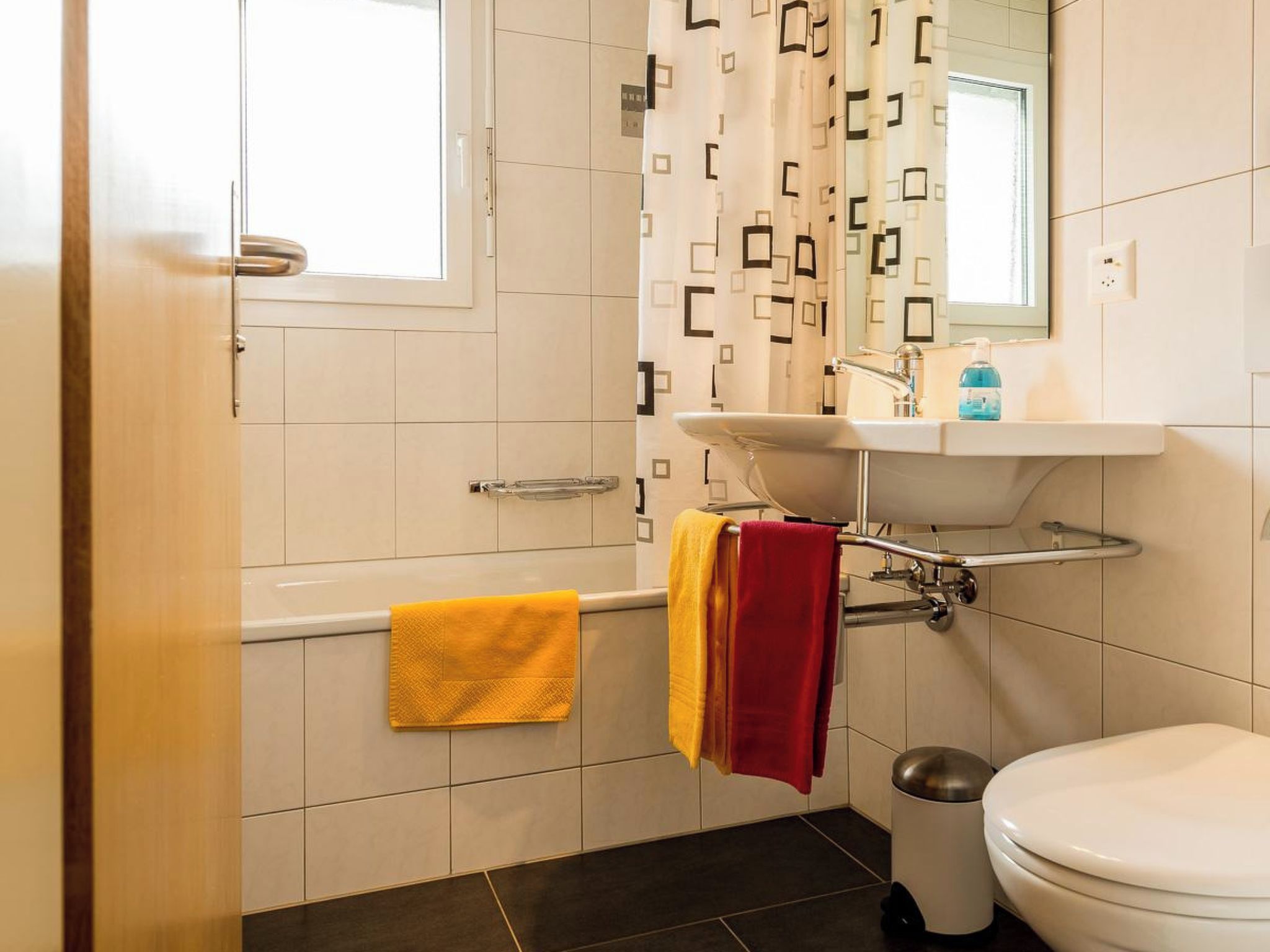 Ett modernt badrum med ett vitt handfat, en toalett och ett badkar. Handdukar hänger på väggen.