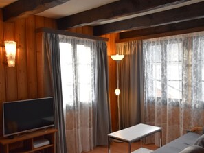 A room with wooden walls, a flat-screen TV on a wooden stand, a table, and two windows with curtains. - Chalet Antika Appartement/Fewo 5-Bettwohnung by Interhome (Saas-Fee)
