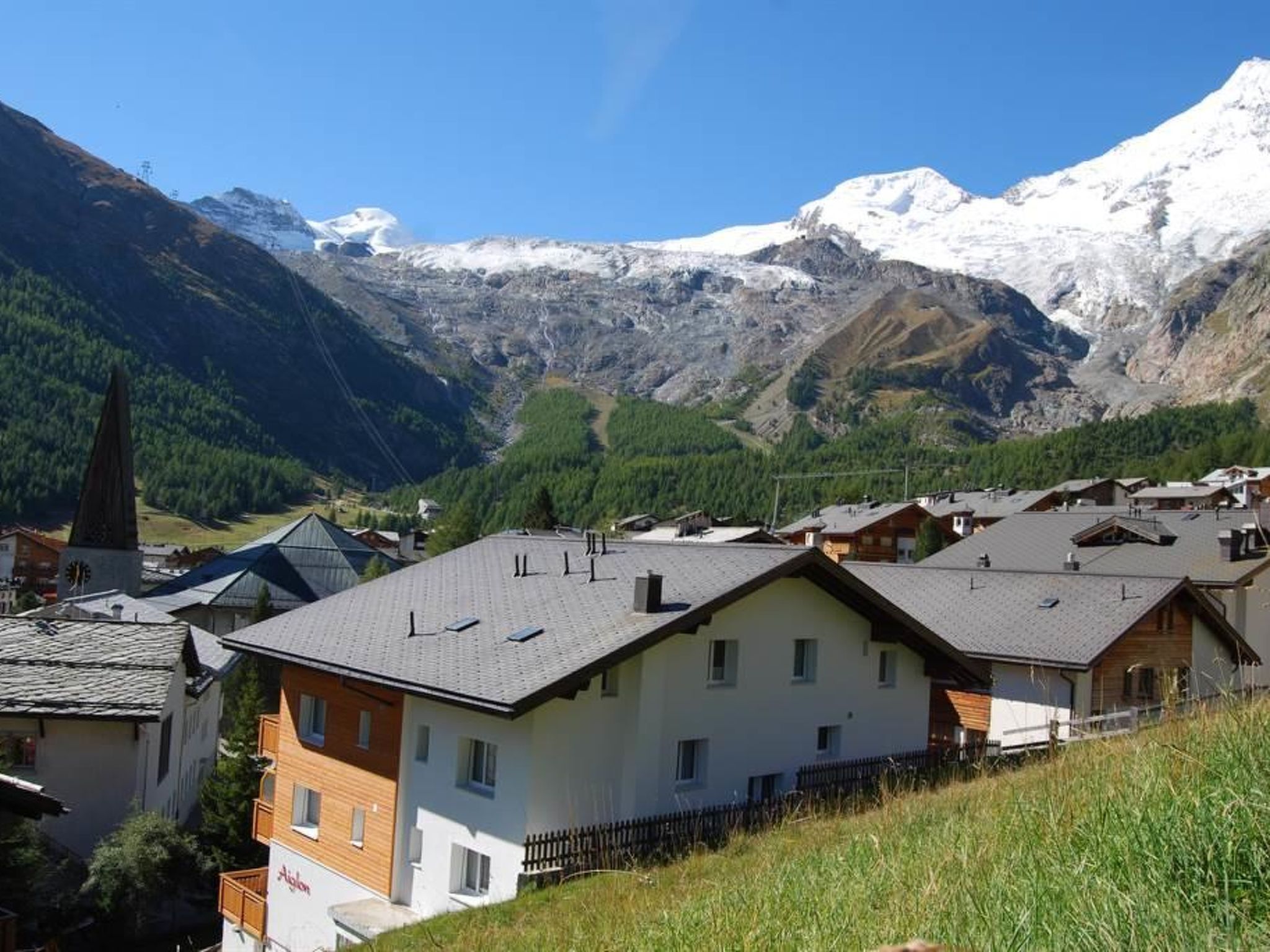 Un pueblo de montaña con casas, una aguja de iglesia y picos nevados al fondo.