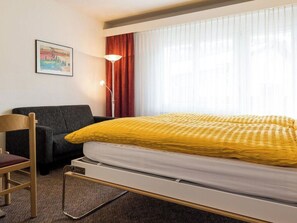 Quarto de hotel com cama, poltrona, escrivaninha, abajur e janela com cortinas.