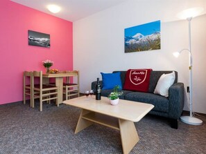 A room with a wooden table, a sofa with a red cushion, a chair, a floor lamp, and two wall-mounted pictures. - Aiglon Appartement/Fewo 2-Bettwohnung Nr. 2 by Interhome (Saas-Fee)