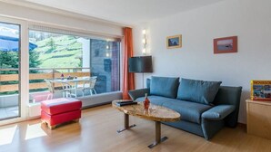 Ett modernt vardagsrum med en soffa, ett litet bord och en röd pall. Det finns ett stort fönster med utsikt över grönområden utanför.
