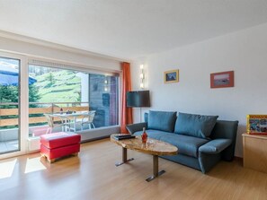 A modern living room with a sofa, a small table, and a red ottoman. There is a large window with a view of greenery outside. - Acimo Appartement/Fewo 2-Bettwohnung Muschi by Interhome (Saas-Fee)