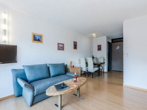 A living room with a blue sofa, a coffee table, a TV mounted on the wall, and a dining area with chairs and a table. - Acimo Appartement/Fewo 2-Bettwohnung Muschi by Interhome (Saas-Fee)