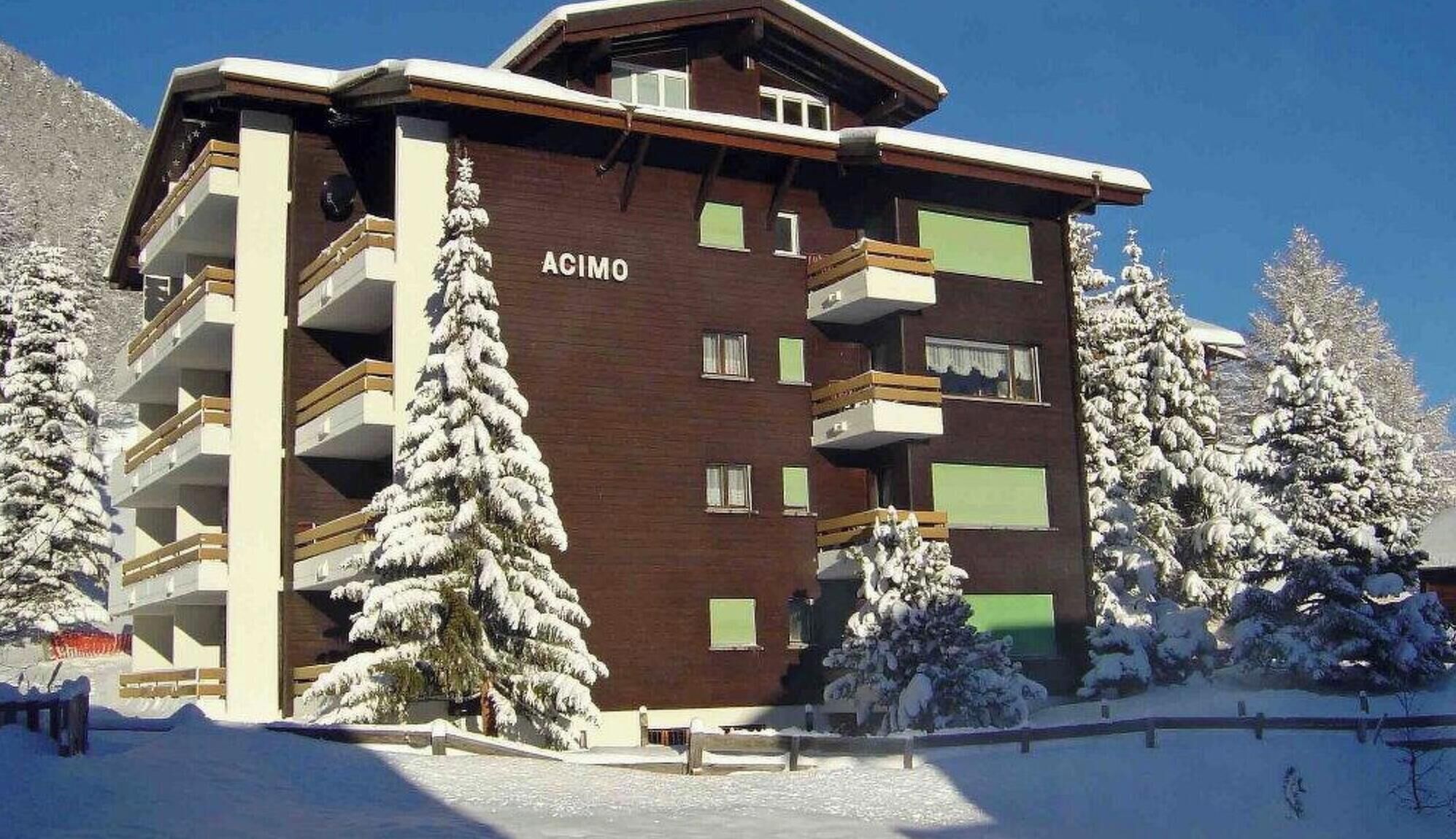 A multi-story building with balconies, labeled 'ACIMO,' surrounded by snow-covered trees.