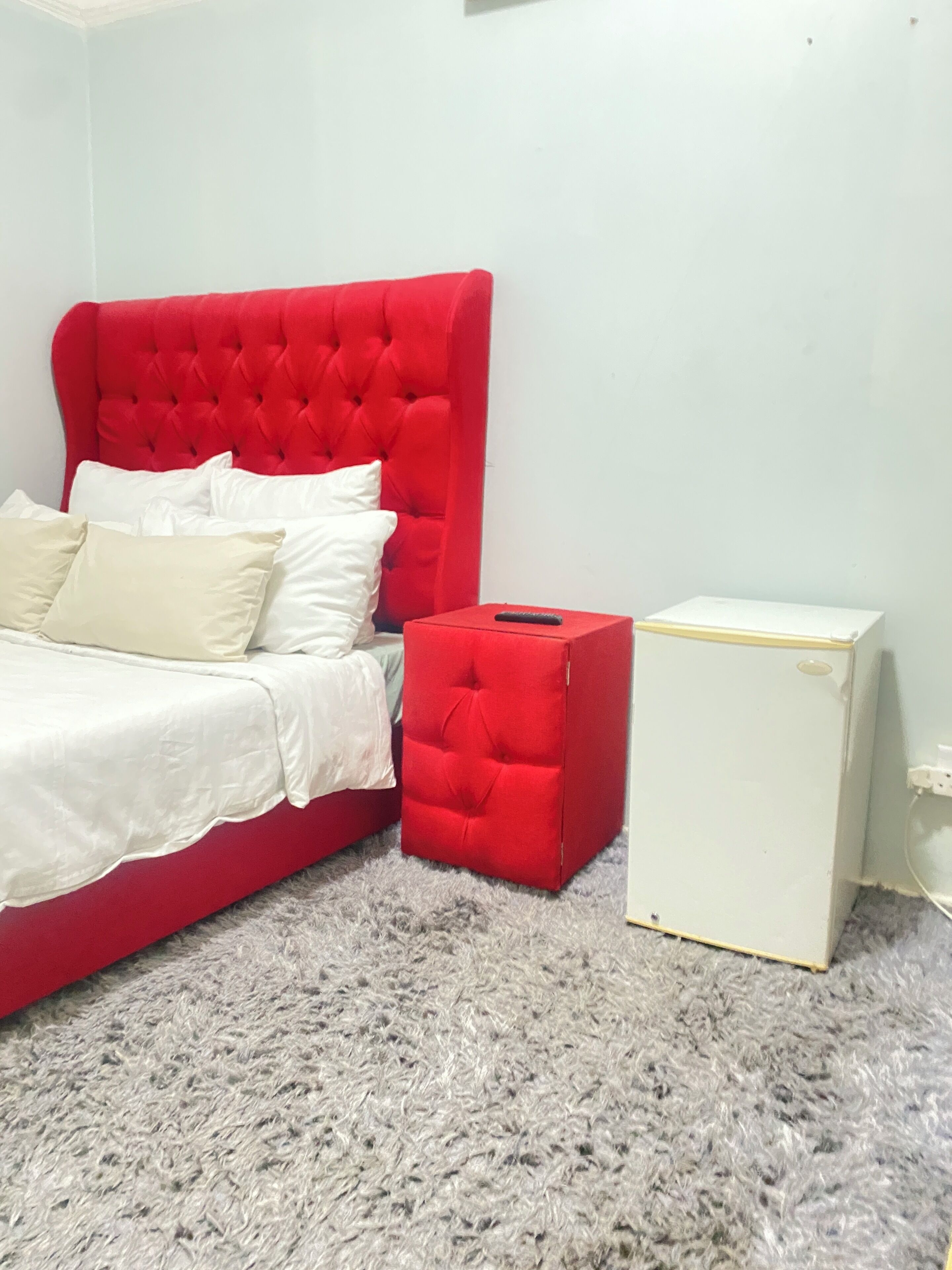A bed with a red headboard, white bedding, and a red ottoman. There is a white bedside table with a remote control on it. The room has a grey carpet.