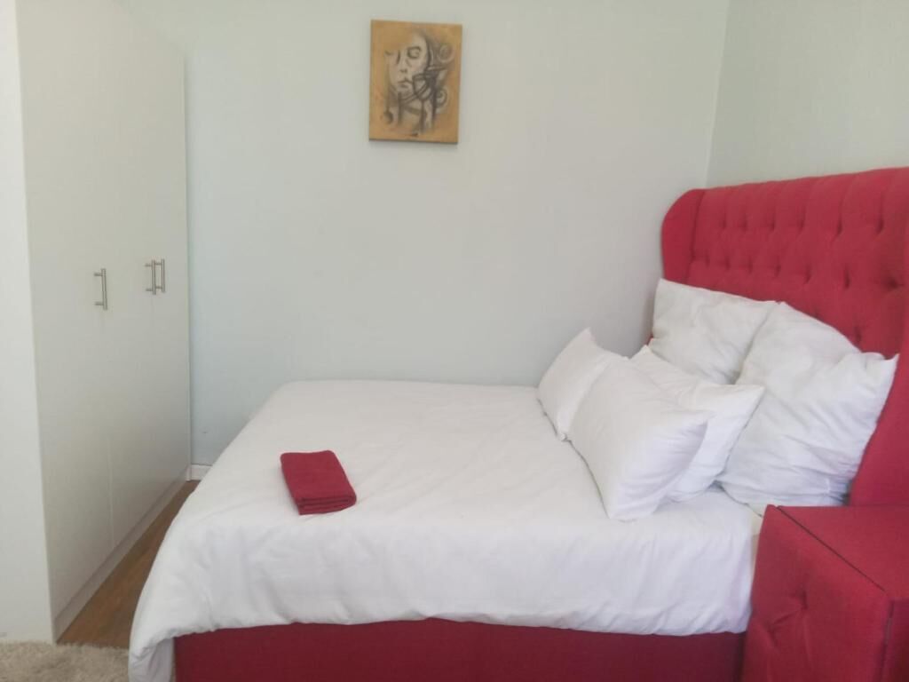 Een bed met witte beddengoed en rode accenten, een rood hoofdeinde en een schilderij aan de muur.
