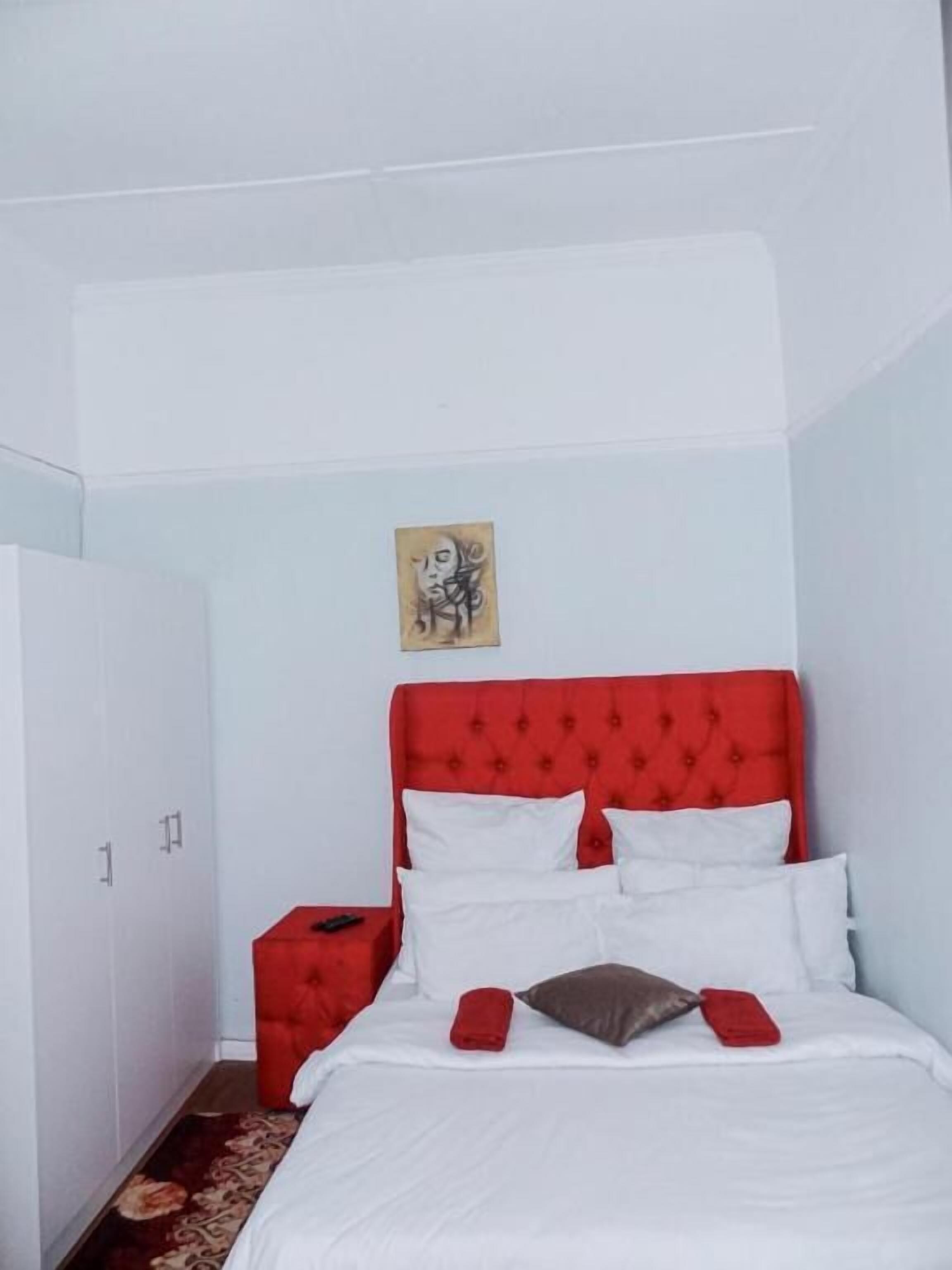A bedroom with a red upholstered headboard, white bedding, a small red nightstand, and a single artwork on the wall.