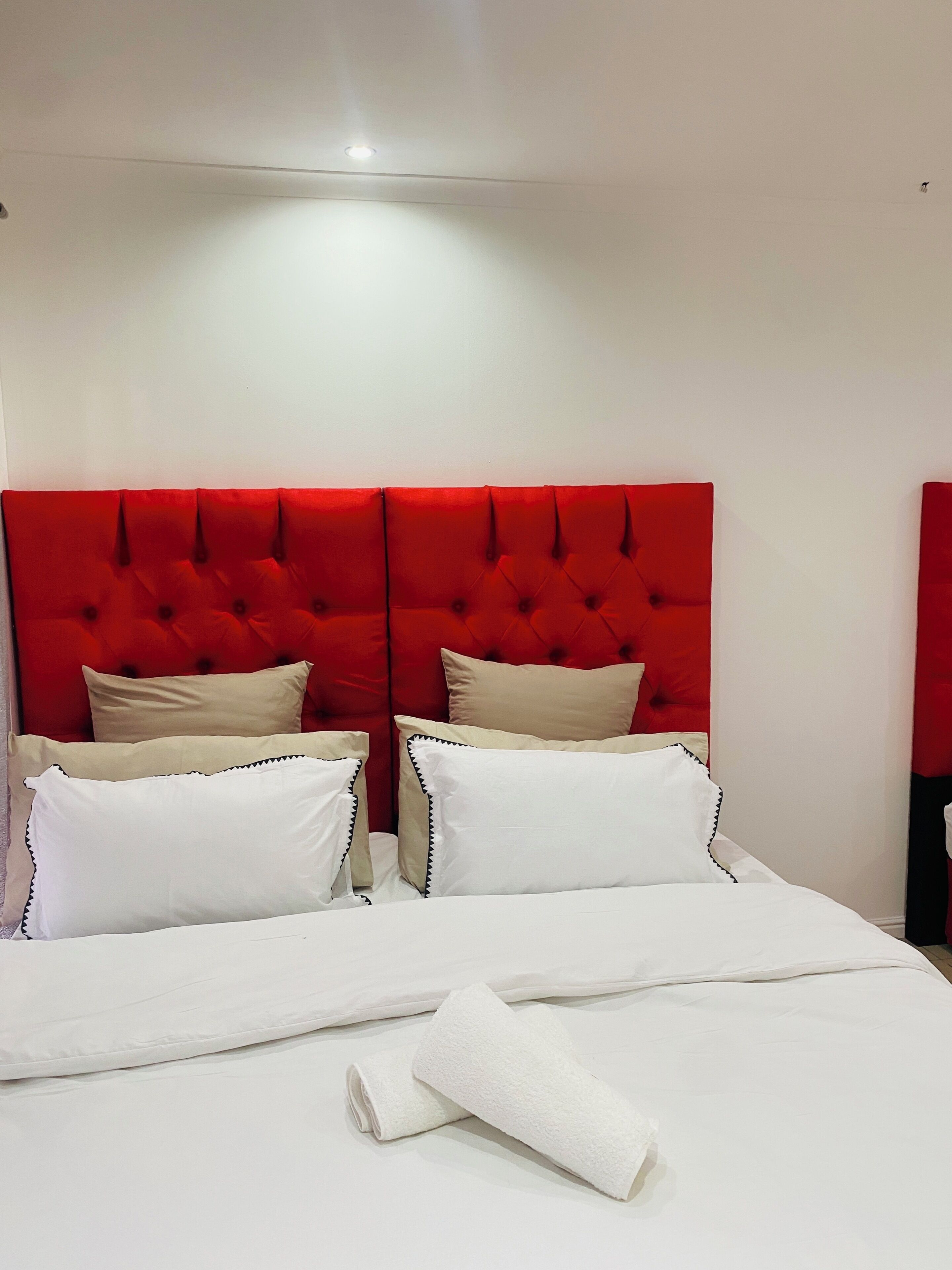 Camera d'albergo con una testiera imbottita rossa, biancheria bianca e un asciugamano piegato con cura sul letto.