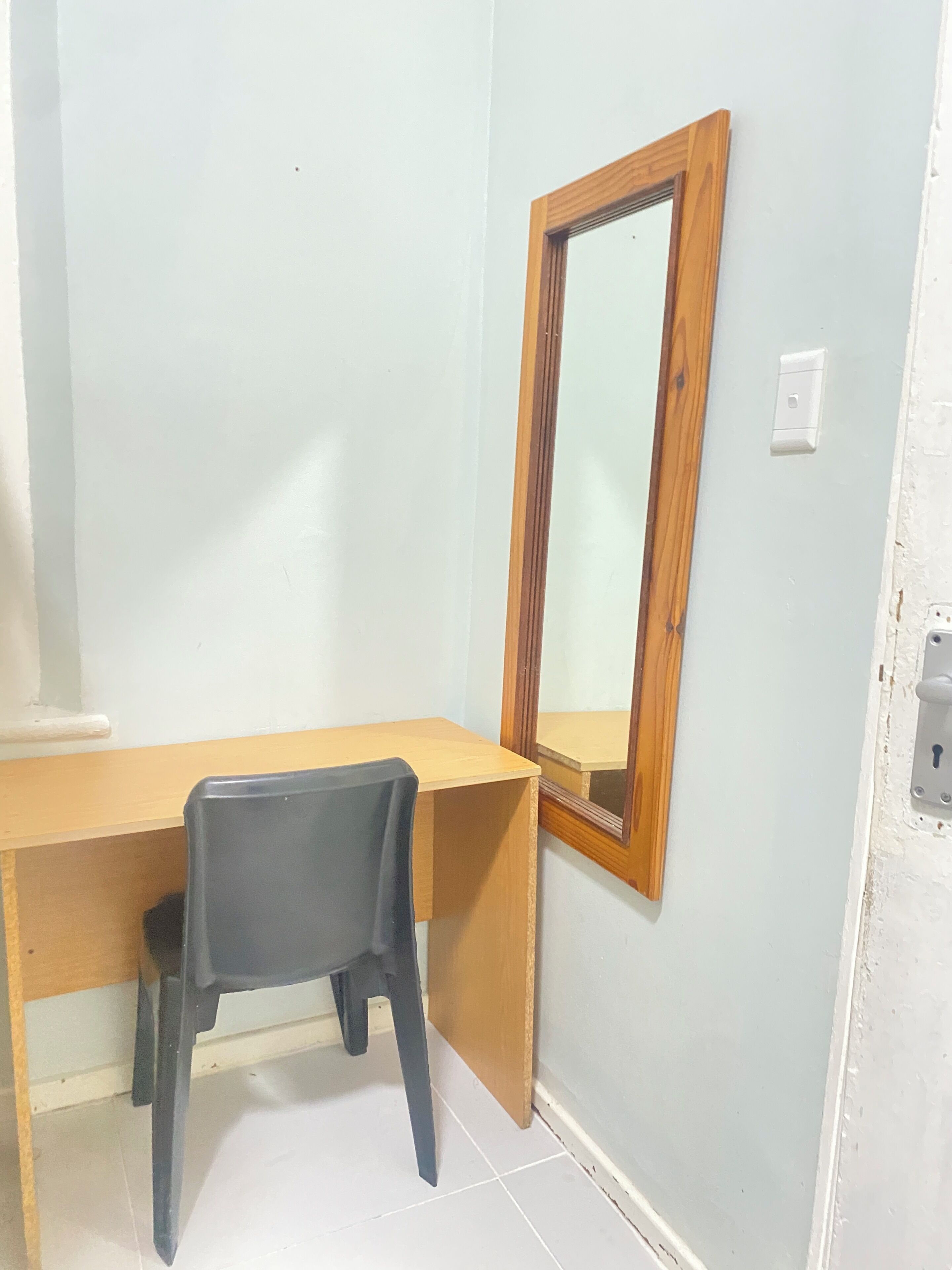 A room with a wooden desk, a black chair, a wooden-framed mirror, and a light switch on the wall.
