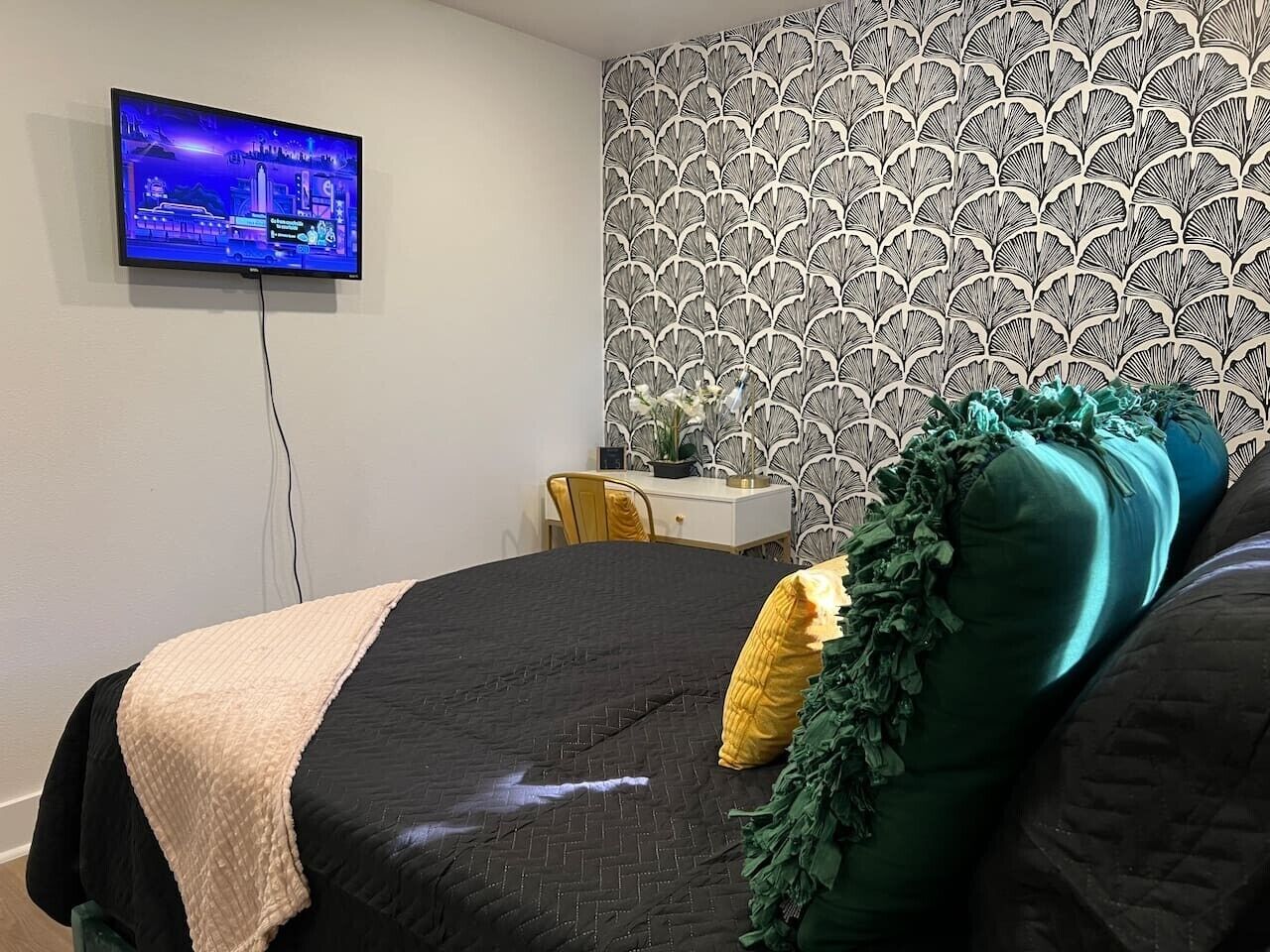 A bedroom with a bed, a TV mounted on the wall, a desk, and a patterned wallpaper.
