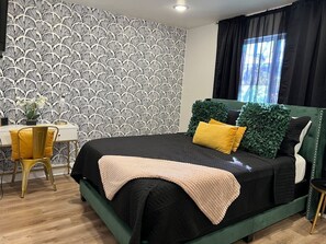 A modern bedroom with a patterned wallpaper, a green bed with black and yellow bedding, a desk with a chair, and a window with black curtains. - Fair Park Area | Free Parking | Slps 4 (Dallas)