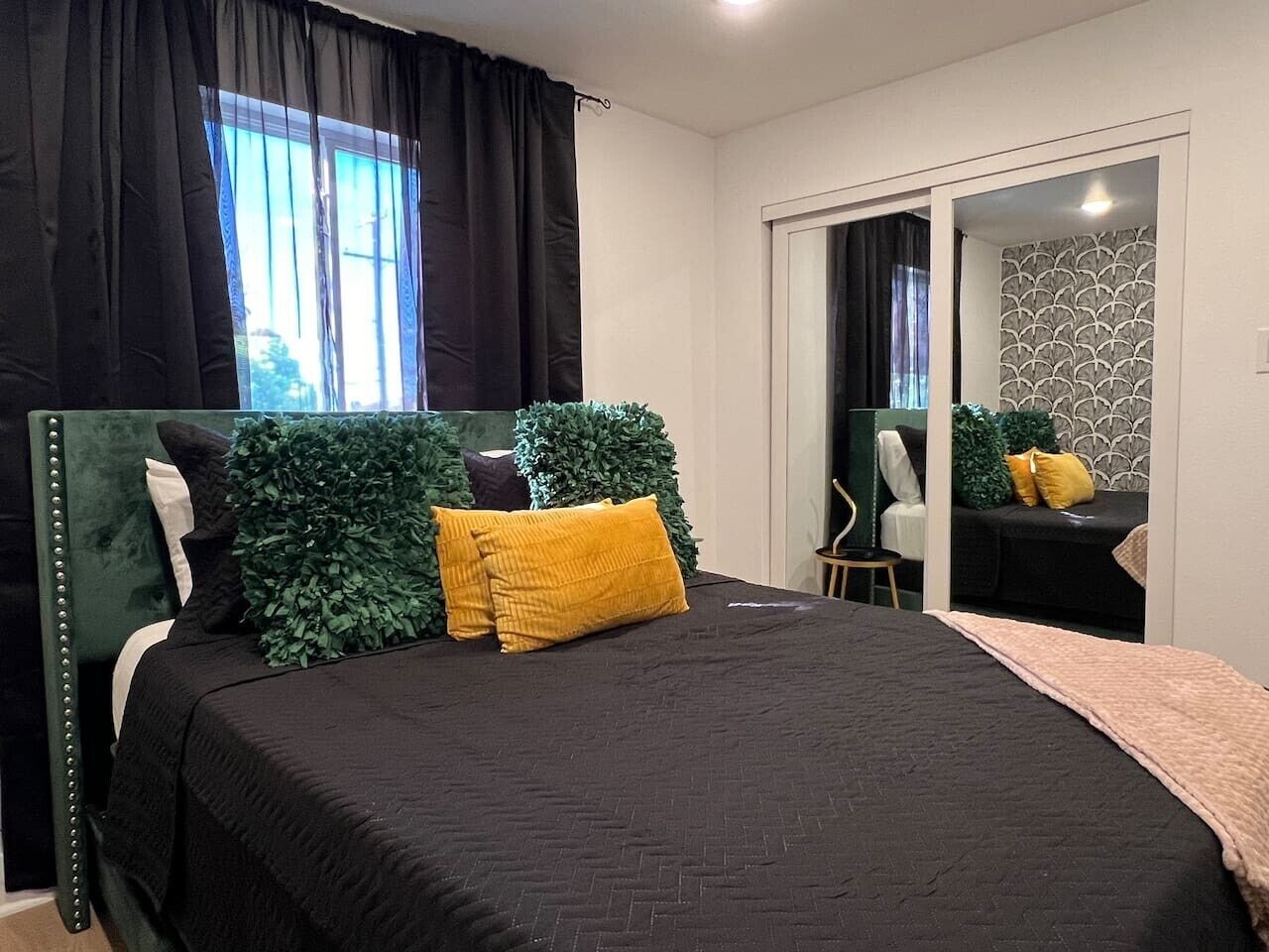 Un dormitorio con cama, espejo, una mesita y ventana con cortinas.