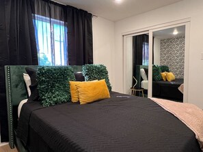 Ein Schlafzimmer mit Bett, Spiegel, einem kleinen Tisch und einem Fenster mit Vorhängen.