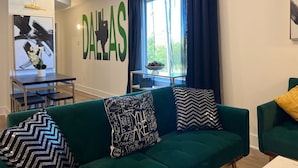Uma sala de estar com um sofá verde, uma mesa de centro, uma televisão e uma janela grande com cortinas.