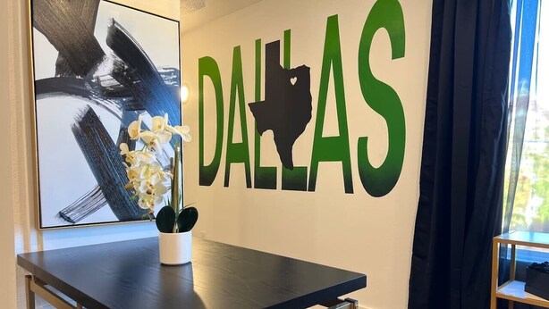 Um quarto com uma mesa, uma parede com a inscrição 'DALLAS', uma obra de arte emoldurada e uma janela com cortinas.