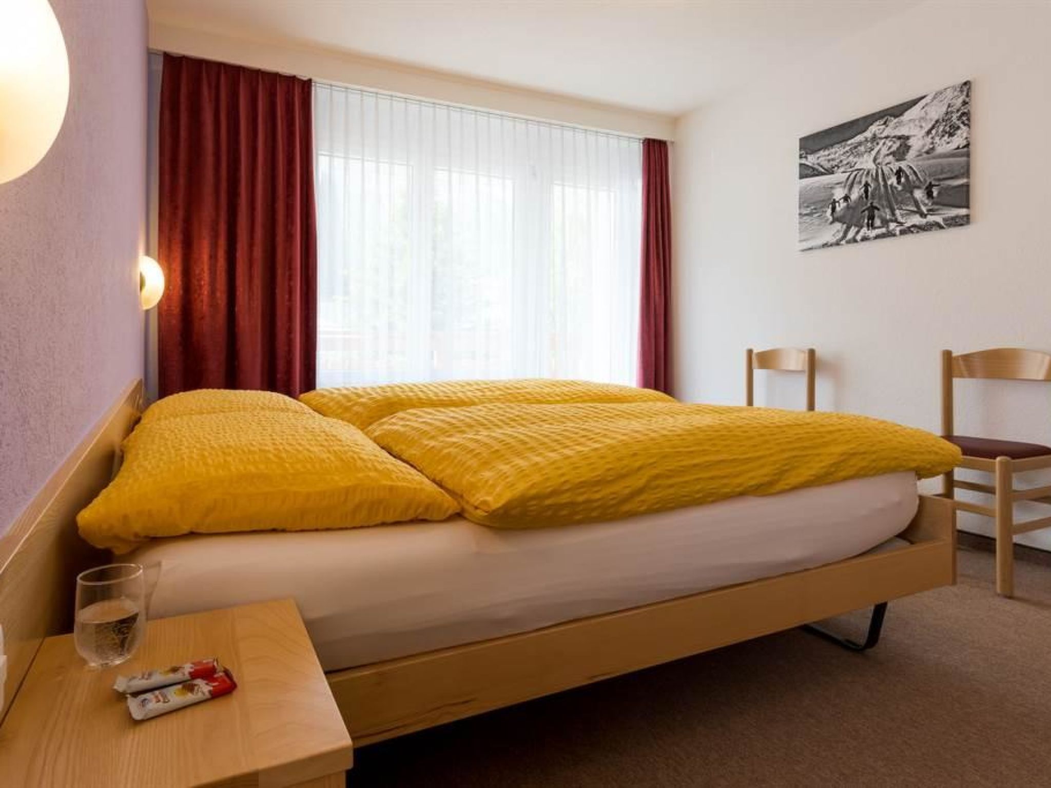 A hotel room with a bed, a bedside table with a glass and a snack, a chair, and a wall-mounted picture.