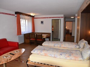 A room with a red sofa, a small table with a remote control, a patterned sofa bed, a dining table with chairs, and a door leading to another room. - Acimo Appartement/Fewo 5-Bettwohnung Romeo & Julia by Interhome (Saas-Fee)