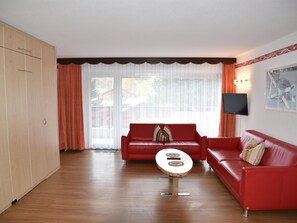 A living room with two red sofas, a coffee table, a TV mounted on the wall, and a large window with curtains. - Acimo Appartement/Fewo 5-Bettwohnung Romeo & Julia by Interhome (Saas-Fee)