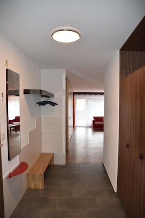 Um corredor com um banco de madeira, um cabideiro e um espelho. Há uma porta à direita e um sofá vermelho ao fundo.