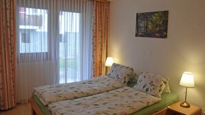 Een tweepersoonsbed met bloemenbeddengoed, twee bedlampjes, een schilderij aan de muur en uitzicht door een raam met dunne gordijnen.