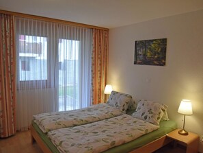 Ein Doppelbett mit floraler Bettwäsche, zwei Nachttischlampen, ein Bild an der Wand und Blick durch ein Fenster mit durchsichtigen Vorhängen.