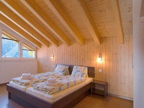 A room with a wooden ceiling, a bed with patterned bedding, two bedside tables, and a window with a view. - Chalet Les Arolles Appartement/Fewo 10-Bettwohnung by Interhome (Saas-Fee)