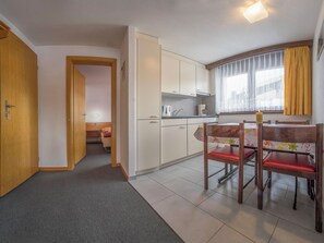 A compact kitchen with white cabinets, a dining area with two wooden chairs, and a window with yellow curtains. - Bergrose Appartement/Fewo 4-Bettwohnung Nr. 1 by Interhome (Saas-Fee)