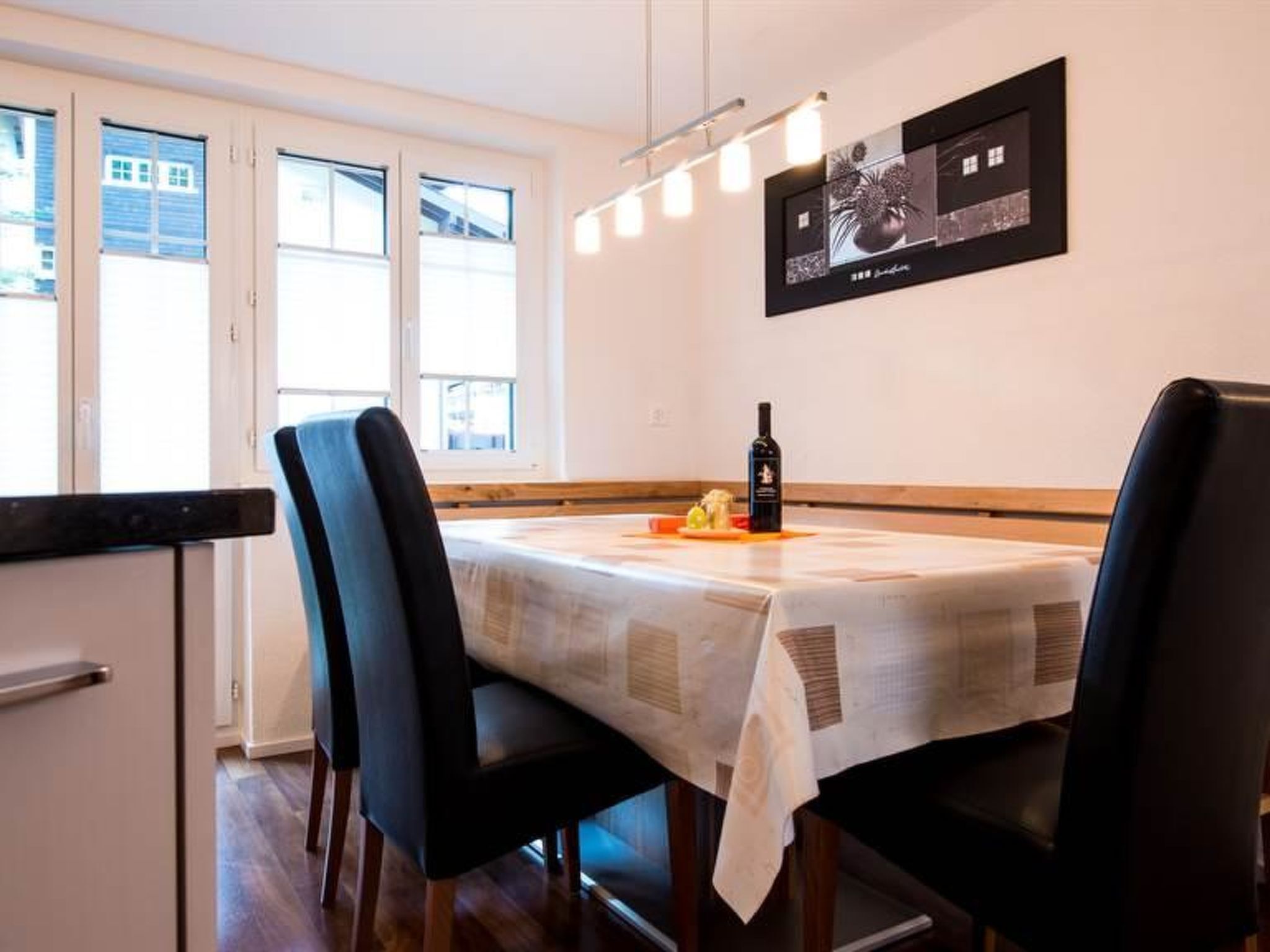 Un coin repas avec une table dressée pour deux personnes, des chaises, une bouteille de vin et une œuvre d’art encadrée accrochée au mur.