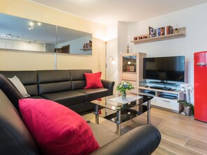Een moderne woonkamer met een zwarte bank, rode kussens, een glazen salontafel, een flatscreen-tv en een rode koelkast.