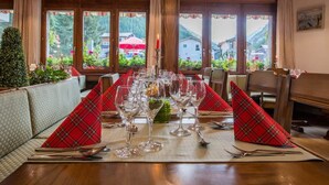 Een restaurant met een lange tafel gedekt voor een maaltijd, houten balken in het plafond en grote ramen met uitzicht op groen en gebouwen.
