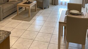 Tilava olohuone, jossa on korkea katto, sohva, sohvapöytä, kahdelle katettu ruokapöytä ja ikkuna, jossa on verhot.