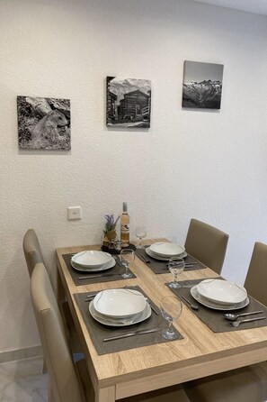 Une table à manger est dressée pour quatre personnes, avec des assiettes, des verres et des couverts, et des chaises sont placées de chaque côté. Trois photographies en noir et blanc sont accrochées au mur au-dessus de la table.