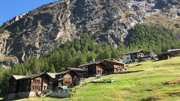 Un pueblo de montaña con casas de madera tradicionales, cielo despejado y una imponente montaña rocosa al fondo.