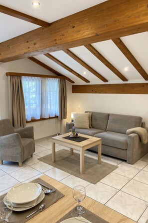 Un salon avec un plafond en bois, un canapé, un fauteuil, une table basse et un coin repas avec une table dressée pour deux personnes.