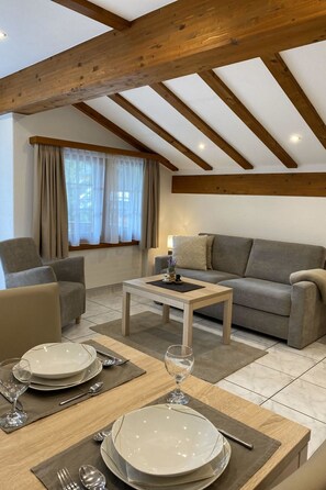 A furnished living room with a sofa, a dining table set for two, and a window with curtains. - Apartments Arcadia Appartement/Fewo 4-Bettwohnung Dom by Interhome (Saas-Grund)