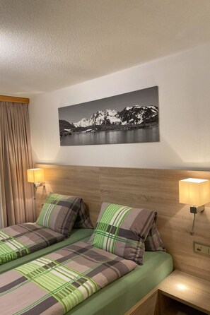 Une chambre d’hôtel avec un lit, deux lampes de chevet, un tableau représentant un paysage de montagne accroché au mur, et une tête de lit en bois.