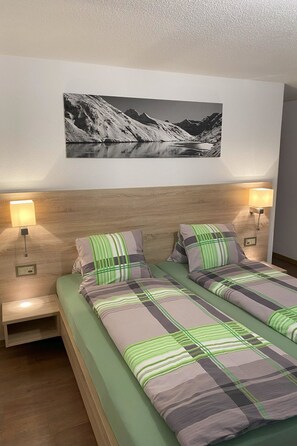A double bed with striped bedding, two bedside lamps, a wall-mounted picture of a mountain landscape, and a wooden headboard. - Apartments Arcadia Appartement/Fewo 6-Bettwohnung Allalin by Interhome (Saas-Grund)