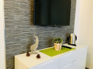 A modern living room with a flat-screen TV mounted on a textured wall, a white cabinet with a green placemat, a decorative wooden goat, a potted plant, and a coffee maker. - Alcazar Apartments Appartement/Fewo Wohnung 2 by Interhome (Saas-Grund)